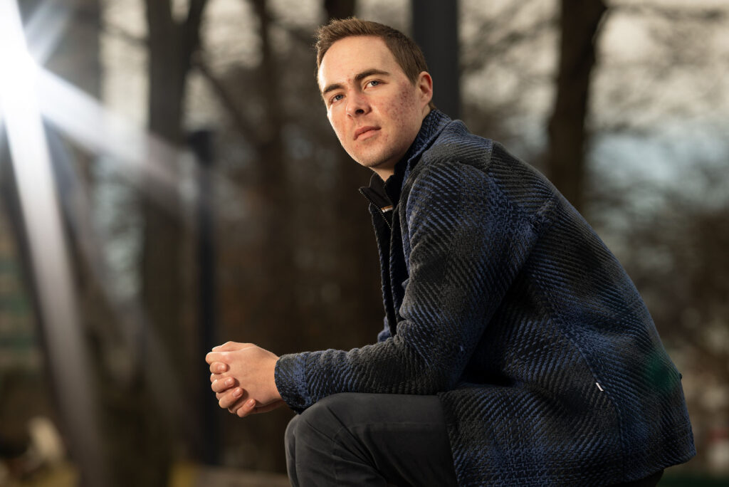 Photo: Cameron Monesmith, a white man with short hair wearing a flannel shirt, sits outside, his left side profile to the camera, hands clasp, posing for the photo.