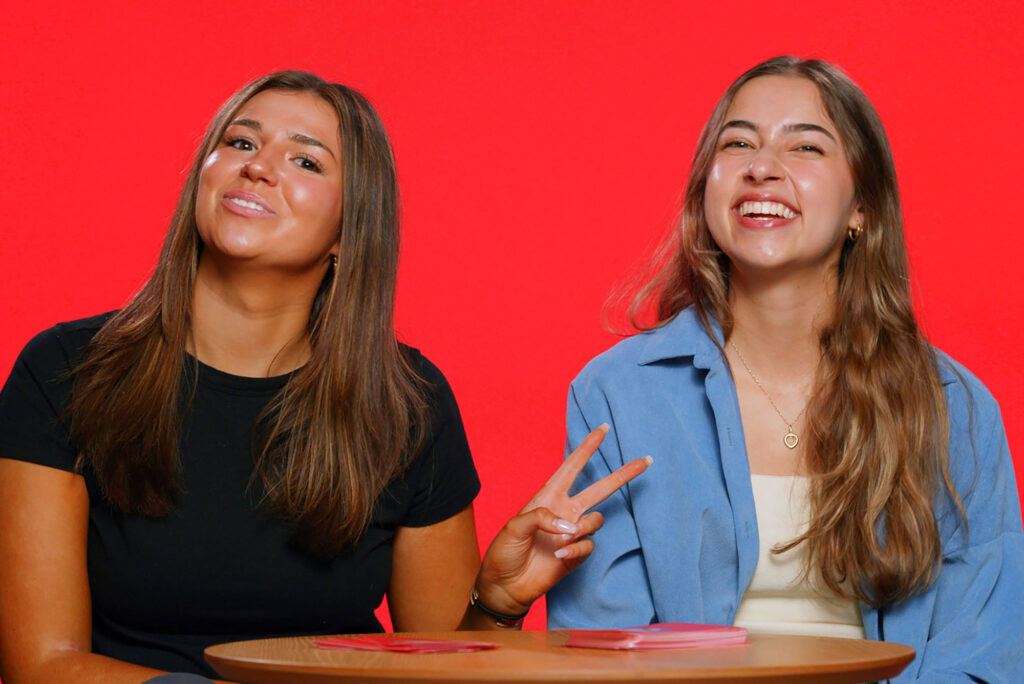 Photo: Two white young women in casual wear smile and pose for the camera in front of a bright red background. They sit at a small, round wooden table with a small pile of red cards on it.