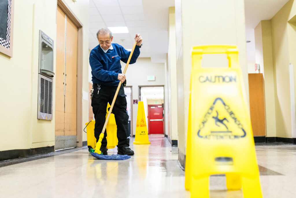 Photo: An older custodian mops a floor in front of a yellow caution slip sign